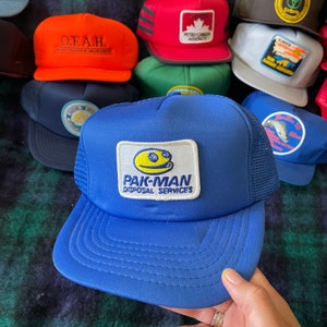 May include: Blue and yellow trucker hat with a smiling face logo and the text "PAK-MAN DISPOSAL SERVICES".
