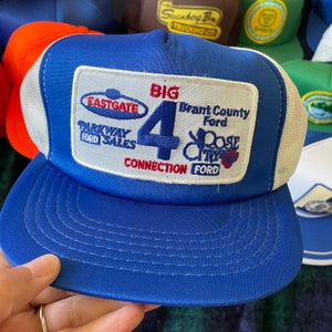 May include: A blue trucker hat with a white mesh back and a curved peak. The front features a rectangular patch with the text "BIG 4 CONNECTION FORD" and other text. The hat is held by a hand.