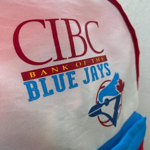 May include: A white backpack with a red and blue CIBC Bank of the Blue Jays logo. The logo features a blue jay with a baseball and a maple leaf.