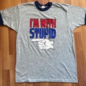 May include: Gray ringer t-shirt with navy blue trim. The shirt features the text "I'M WITH STUPID" in red and blue, with a pointing hand graphic. The shirt is a vintage style.