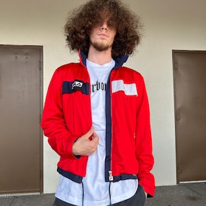 Könnte beinhalten: Eine rot-blaue Windbreaker-Jacke mit einem weißen Patch auf der linken Brust, auf dem in Schwarz "LA" steht. Die Jacke hat einen Reißverschluss und einen Kordelzug am unteren Rand.