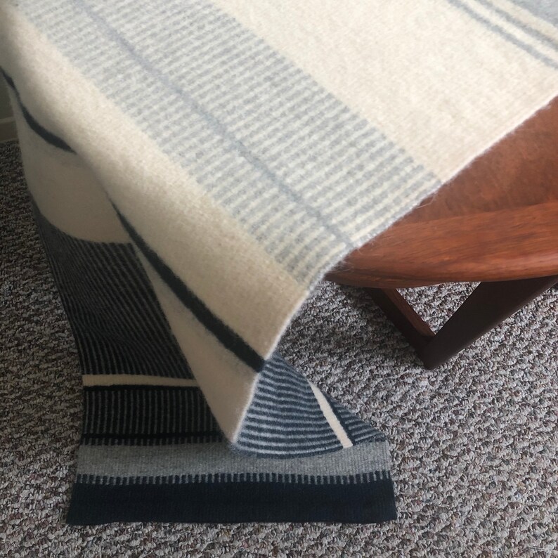 May include: A white and gray striped throw blanket with a black and gray striped border. The blanket is folded and draped over a wooden table.