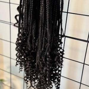 Può includere: Primo piano di trecce marrone scuro con estremità ricci. Le trecce sono appese su uno sfondo a griglia nera. Le trecce sono intrecciate fitte e hanno un aspetto naturale.