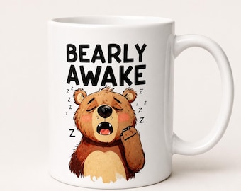 Op de afbeelding: Een witte keramische mok met een illustratie van een bruine beer met gesloten ogen en een open mond, die lijkt te gapen. De tekst "BEARLY AWAKE" staat in zwarte hoofdletters boven de beer, vergezeld door kleine zwarte "Z" symbolen.