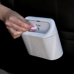 Pode incluir: Um pequeno caixote do lixo branco e angular para carro, com tampa de botão. A tampa tem a palavra "PUSH" e um ícone de mão. O caixote está colocado contra um banco de carro preto.
