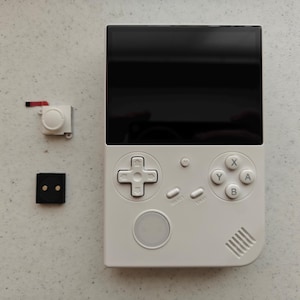 May include: A white handheld gaming console with a black screen, D-pad, and action buttons. A white joystick and a black square component are placed next to the console. The console has a speaker and select/start buttons.