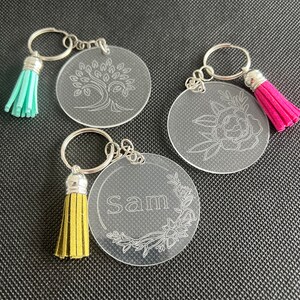 K&ouml;nnte beinhalten: Drei transparente Acryl-Schl&uuml;sselanh&auml;nger mit silbernen Schl&uuml;sselringen und farbigen Quasten. Ein Schl&uuml;sselanh&auml;nger zeigt ein Baumdesign, ein weiterer ein Blumendesign und der dritte den Namen "Sam" mit floralen Akzenten. Quasten sind t&uuml;rkis, rosa und gelb.