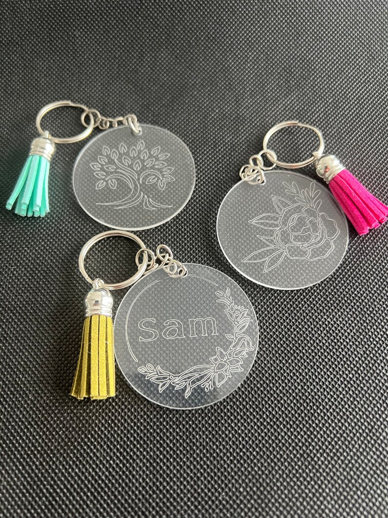 K&ouml;nnte beinhalten: Drei transparente Acryl-Schl&uuml;sselanh&auml;nger mit silbernen Schl&uuml;sselringen und farbigen Quasten. Ein Schl&uuml;sselanh&auml;nger zeigt ein Baumdesign, ein weiterer ein Blumendesign und der dritte den Namen "Sam" mit floralen Akzenten. Quasten sind t&uuml;rkis, rosa und gelb.