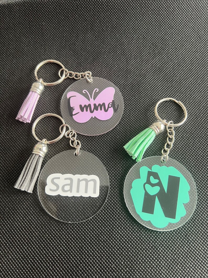 K&ouml;nnte beinhalten: Drei transparente Acryl-Schl&uuml;sselanh&auml;nger mit silbernen Schl&uuml;sselringen und Quasten. Einer zeigt ein lila Schmetterlingsdesign mit dem Namen "Emma". Ein anderer hat den Namen "Sam" in Wei&szlig;. Der dritte hat ein gr&uuml;nes Design mit dem Buchstaben "N".