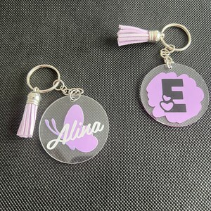 K&ouml;nnte beinhalten: Zwei Schl&uuml;sselanh&auml;nger aus klarem Acryl mit silberfarbener Hardware und lavendelfarbenen Quasten. Einer zeigt einen lavendelfarbenen Schmetterling und den Namen "Alina" in wei&szlig;er Schrift. Der andere hat einen lavendelfarbenen Hintergrund mit dem Buchstaben "E" und zwei Herzen.