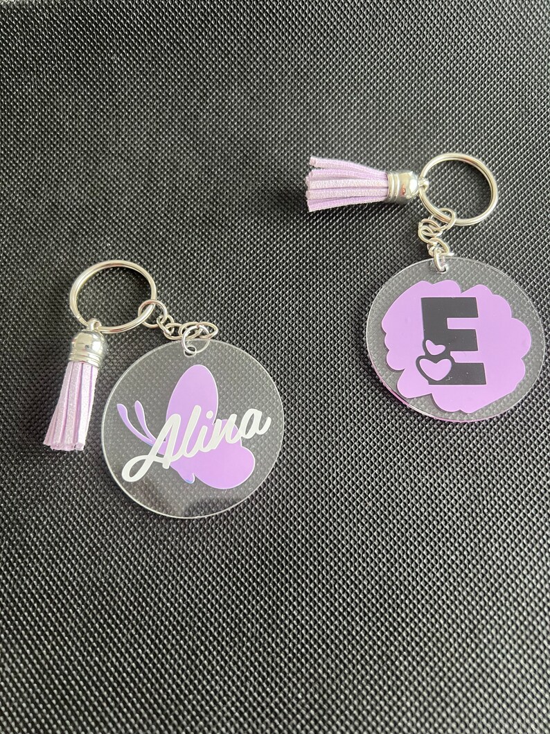 K&ouml;nnte beinhalten: Zwei Schl&uuml;sselanh&auml;nger aus klarem Acryl mit silberfarbener Hardware und lavendelfarbenen Quasten. Einer zeigt einen lavendelfarbenen Schmetterling und den Namen "Alina" in wei&szlig;er Schrift. Der andere hat einen lavendelfarbenen Hintergrund mit dem Buchstaben "E" und zwei Herzen.