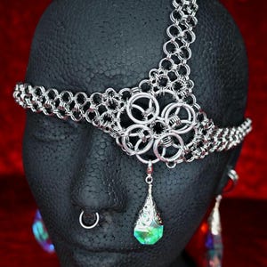 Puede incluir: Una diadema de cota de malla plateada con un diseño floral central y un colgante en forma de lágrima con un cristal verde y azul. La diadema se muestra sobre una cabeza de maniquí negra sobre un fondo rojo.