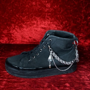 Può includere: Sneaker alta nera con suola spessa. La scarpa presenta dettagli con catena e ciondoli argentati, tra cui un ciondolo a forma di teschio. I lacci sono neri, su sfondo rosso.