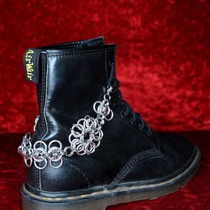 Pode incluir: Bota de couro preta com tornozeleira de corrente prateada. A tornozeleira apresenta um design floral e é presa à bota com uma pequena corrente. A bota tem costuras amarelas e as palavras "Air Wair" impressas nas costas.