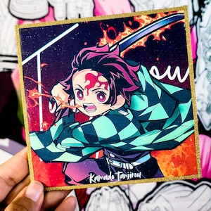 May include: Square sticker featuring an anime character with red hair and a checkered green and black patterned jacket. The character is holding a sword with flames in the background. The sticker has a gold glitter border and the name "Kamado Tanjirou" is written on the bottom.