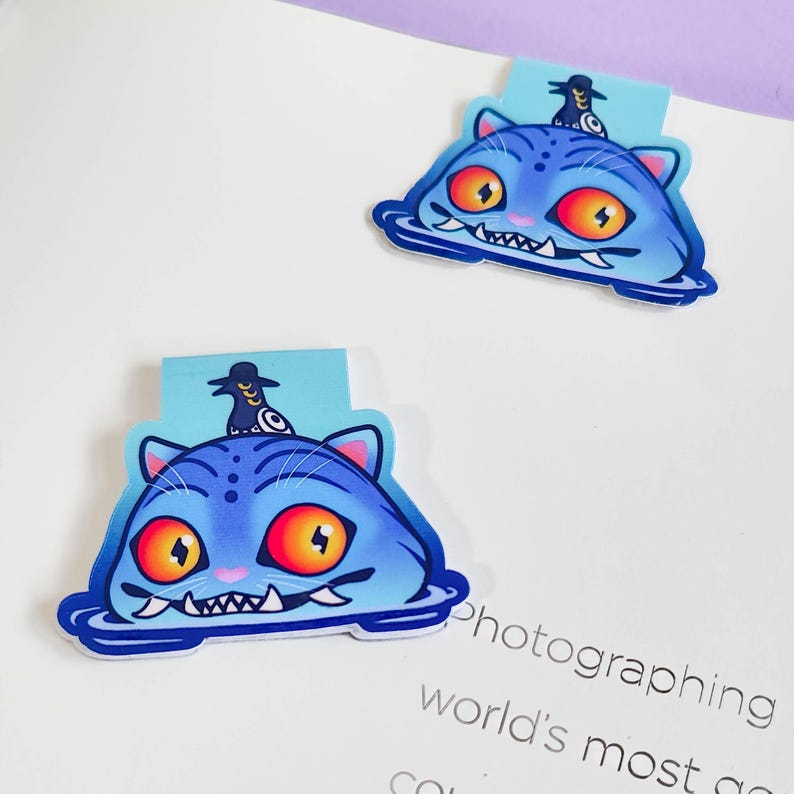 May include: Two blue cat-shaped bookmarks with orange eyes and white teeth. Each cat has a small figure on top. The cats appear to be floating in water. The bookmarks are on a white surface.