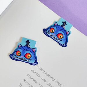 May include: Two blue cat-shaped bookmarks with orange eyes and white teeth are displayed on a white surface. Each bookmark has a light blue tab with a black figure on top. The bookmarks are placed on a book page.