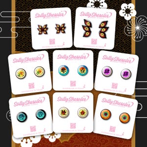 May include: A collection of handmade stud earrings with various designs. The earrings feature colorful graphics, including abstract shapes and Japanese characters. Each pair is displayed on a white card with the text "Sally Shesales GOODY STUDIO HANDMADE IN TEXAS".