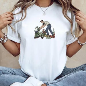May include: White t-shirt featuring a vintage-style illustration of a person tending a garden. The graphic depicts a person in a white shirt and blue pants among various plants and vegetables. The design has a retro aesthetic.
