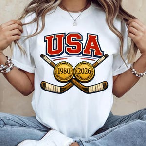 May include: White t-shirt with the word "USA" in red and blue block letters. Below the text are two crossed hockey sticks and two gold medallions with the years "1980" and "2026".