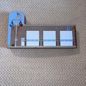 Puede incluir: Un modelo de madera de una estación de bomberos con tres puertas de garaje, una puerta pequeña y un asta de bandera con una bandera española y una bandera del departamento de bomberos. El edificio está pintado de marrón con molduras blancas y puertas de garaje azules.