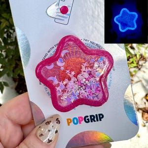 Może przedstawiać: Różowy PopGrip w kształcie gwiazdy z błyszczącą obwódką, wypełniony konfetti o tematyce niebieskiej. PopGrip ma przezroczysty środek z księżycami, gwiazdami i koralikami. Na opakowaniu widnieje napis "TRY ME! PULL" i "POPGRIP". Świecąca niebieska gwiazda jest w rogu.