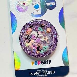 May include: A purple PopGrip with a glittery, celestial design. The round phone accessory features small stars, moons, and other shapes in pink, white, and blue. The packaging reads "PopGrip" and "made with plant-based materials."