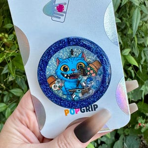 May include: A blue PopGrip featuring a cartoon blue cat character with large yellow eyes, holding a flower pot and a science beaker. The PopGrip has a glittery blue border and the word "PopGrip" is printed in orange and white.
