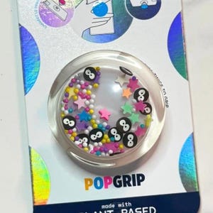 May include: A clear PopGrip with a circular design, filled with colorful beads, stars, and black number eight symbols. The PopGrip is on a white and iridescent background with the word "PopGrip" in yellow and blue.