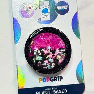 Peut inclure: Un accessoire de téléphone PopGrip avec une bordure noire pailletée et un intérieur rose pailleté. L'intérieur contient des confettis en forme d'étoiles et de chiffres. L'emballage est blanc avec des accents holographiques et le logo PopGrip.