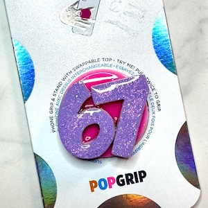 May include: A purple glittery PopGrip phone stand with the number 67. The phone accessory is on a white and silver background with the words "PHONE GRIP & STAND WITH SWAPPABLE TOP" and "POPGRIP" printed on it.