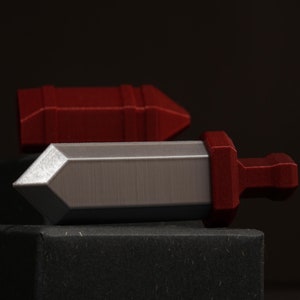 May include: A silver and red plastic sword-shaped object on a black surface. The sword is lying on its side with the tip pointing to the right.