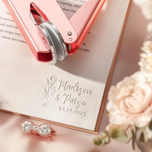 May include: A rose gold embossing machine rests on a cream-colored card, with the embossed names "Madison & Paige" and the date "04.12.2025". A double-stone ring and flowers are also in the image.