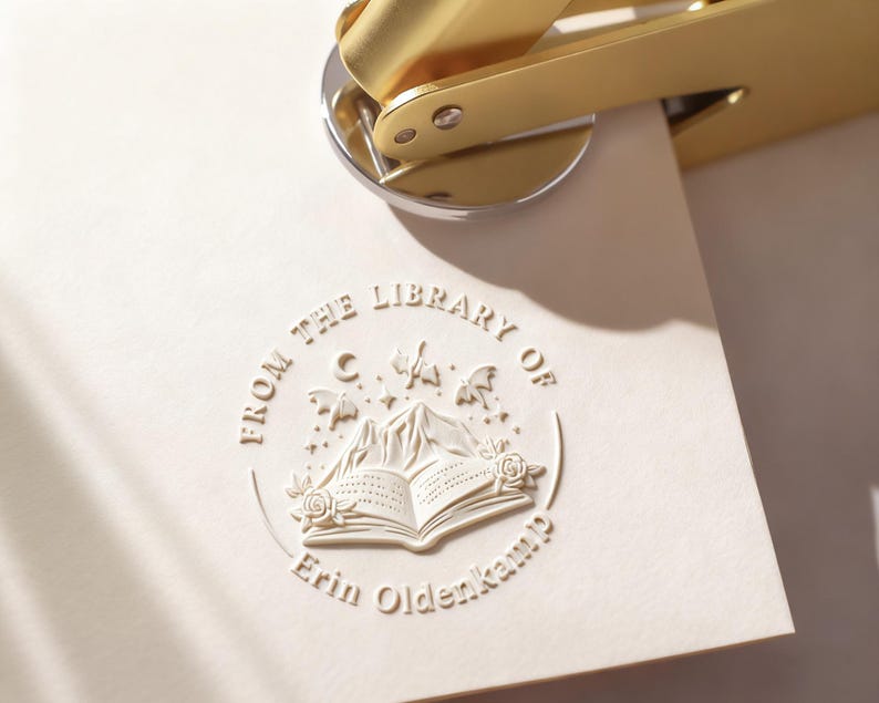 May include: A gold-colored embosser presses a cream-colored paper, creating a raised seal. The seal features an open book, mountains, stars, and the text "FROM THE LIBRARY OF Erin Oldenkamp."