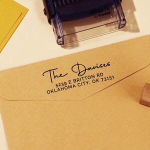 Pode incluir: Um envelope bege com o endereço "The Davises" e seu endereço impresso em tinta preta. Um carimbo preto e transparente está posicionado acima do envelope. O carimbo diz "printy 4913 by trodat".