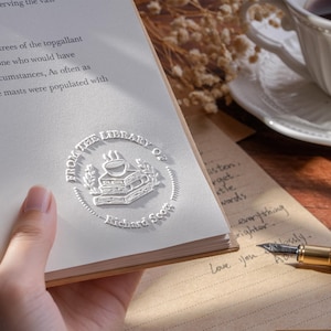 May include: A close-up of a book with a white embossed seal that reads "From the Library of Richard Scott". The book is open, revealing text. A handwritten note and a pen are also visible.