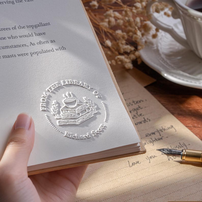 May include: A close-up of a book with a white embossed seal that reads "From the Library of Richard Scott". The book is open, revealing text. A handwritten note and a pen are also visible.