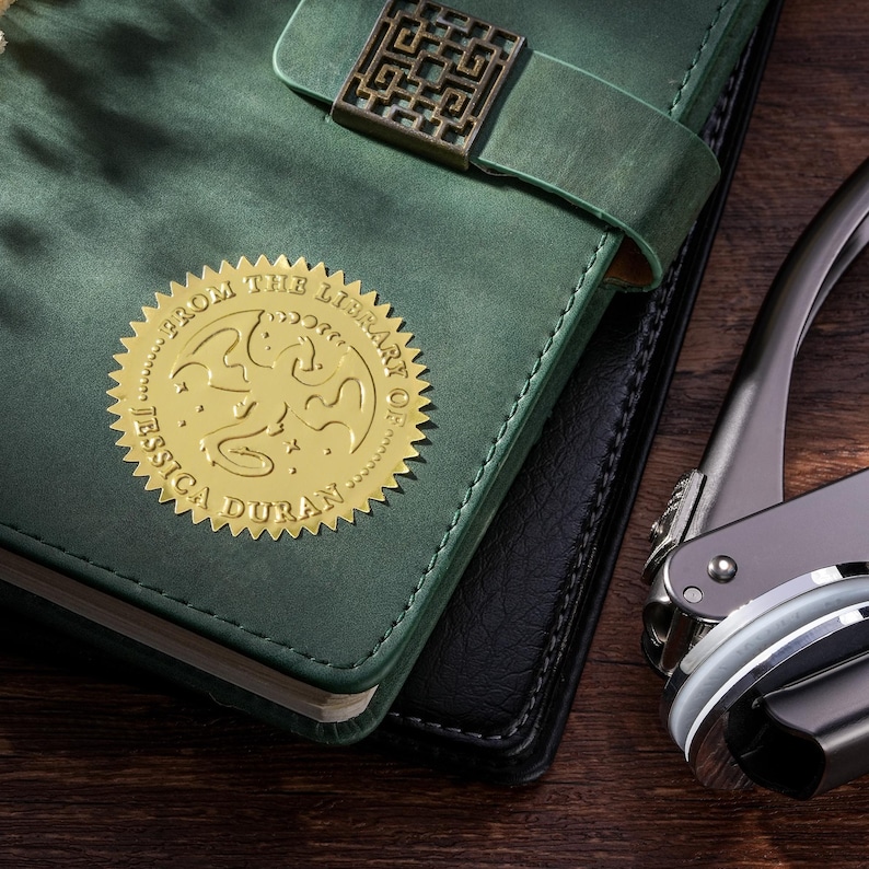 May include: A dark green leather journal with a gold seal that reads "From the Library of Jessica Duran." The journal has a decorative metal clasp. A black leather journal is partially visible beneath the green journal. A silver and black seal press is on the right.