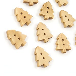 May include: Ten wooden buttons shaped like Christmas trees. Each button is a light brown colour and has two small holes for sewing. The buttons are scattered on a white surface, ideal for craft projects.