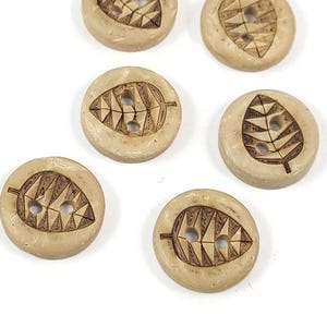 May include: Six round, light tan buttons with a leaf design carved into the surface. Each button has two holes for sewing. The leaf design is a dark brown color, and the buttons are arranged on a white background.