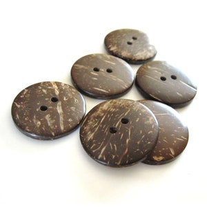 May include: Six brown coconut shell buttons with two holes each. The buttons are arranged in a cluster on a white background.