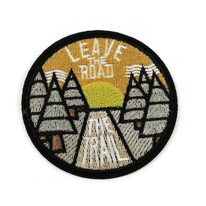 May include: A round, black embroidered patch with a yellow and white design. The patch features a stylised image of a road leading into a forest with a sunset in the background. The text "Leave the Road Take the Trail" is embroidered in white on the patch.