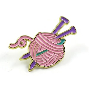 May include: A pink ball of yarn with two purple knitting needles. The yarn ball is gold-coloured and the needles are purple with gold tips.