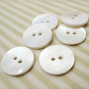 May include: A collection of six round, white buttons with two holes each. The buttons have a pearlescent sheen and are arranged on a striped, beige background. These buttons are suitable for sewing and craft projects.