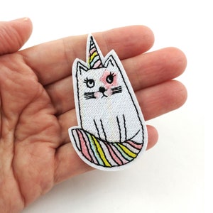 May include: White embroidered iron-on patch of a cat with a pink and yellow unicorn horn and a rainbow striped tail.