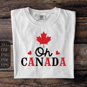 Puede incluir: Camiseta blanca con una hoja de arce roja y el texto "Oh Canada" en negro con dos corazones rojos.