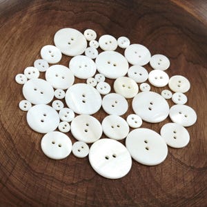 Peut inclure: Un assortiment de boutons blancs ronds, avec deux ou quatre trous, de différentes tailles. Les boutons sont disposés sur une surface en bois brun foncé, créant un effet visuel. Idéal pour la couture et les projets d'artisanat.