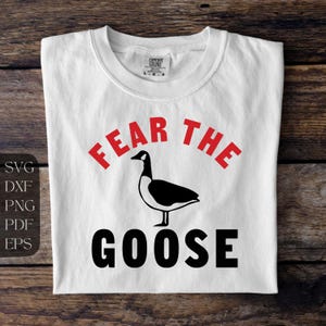 以下が含まれることがあります： 赤と黒の文字で「FEAR THE GOOSE」と書かれた白いTシャツ。黒と白のガチョウのグラフィックが言葉の下に配置されています。木製の表面に折りたたまれたシャツ。