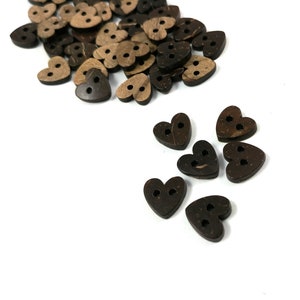 May include: A pile of brown coconut shell heart-shaped buttons with two holes each. There are a few buttons scattered on a white surface.