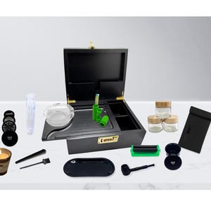 May include: A black wooden box with an open lid, containing various accessories. Includes a grinder, glass jars, a rolling machine, a tray, and a candle. The box has a combination lock and gold-colored hardware.